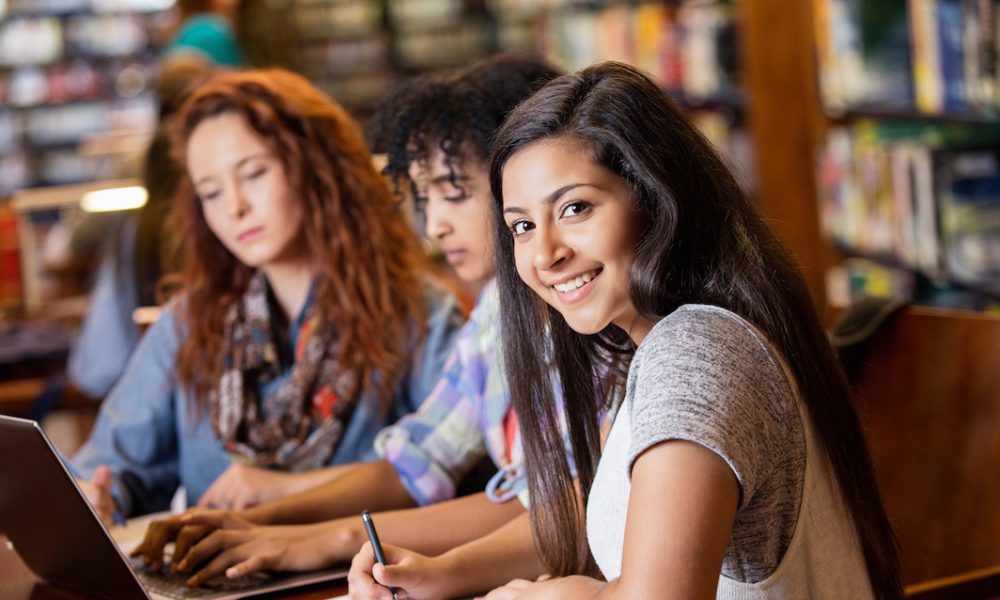 Indian, African American, and Caucasian teenage women are high school or college students. They are sitting at a desk in a crowded library and studying using a laptop computer. Indian girl is smiling and looking at the camera.